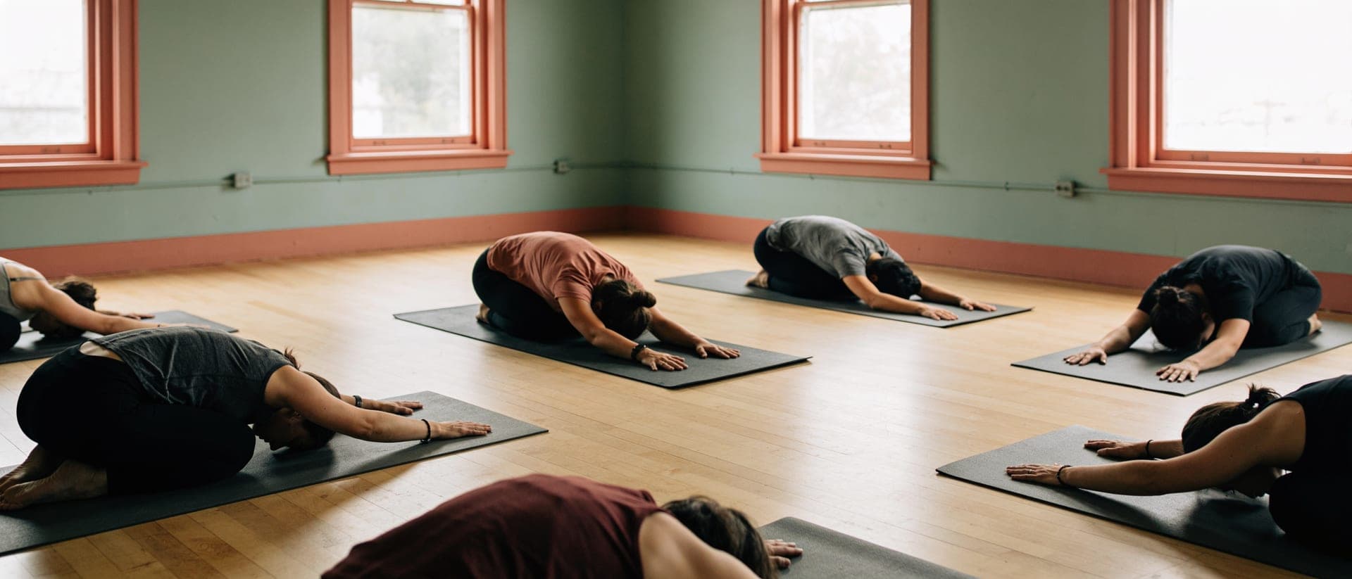 A small group resting in a calm forward fold on mats in a bright, sunlit movement studio.