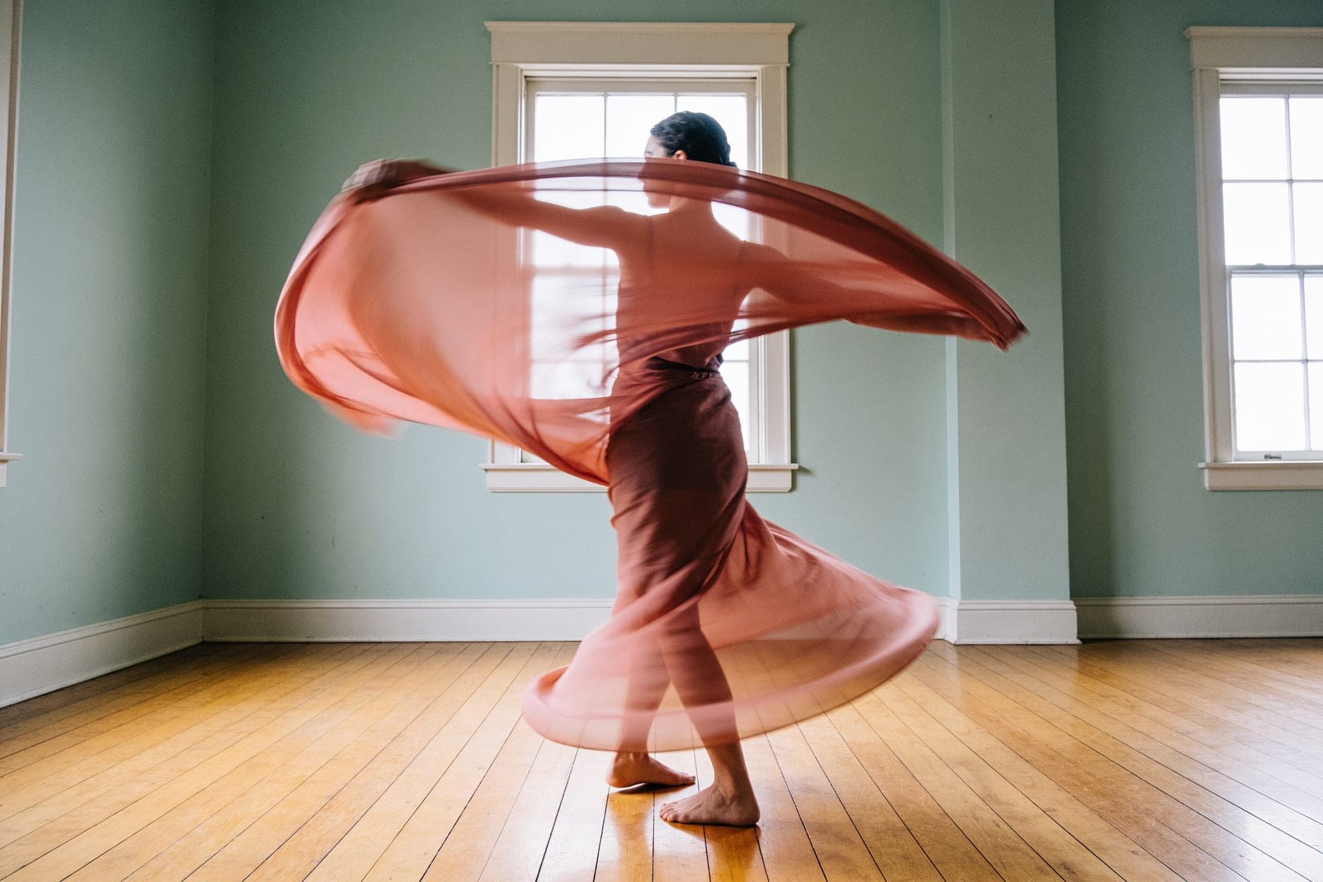 A dancer mid-movement, a sweep of clay-coral fabric trailing in motion across a sunlit wood floor.